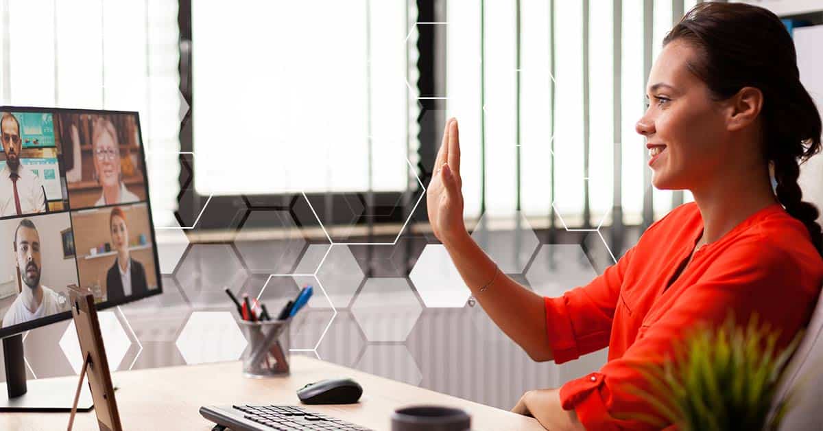 A woman sitting at her desk waving hello at 4 four businesspeople on her screen